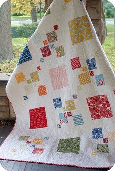 a white quilt with colorful squares on it sitting on top of a wooden bench in front of a tree