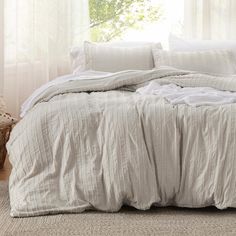 an unmade bed with white linens in front of a window and a basket on the floor