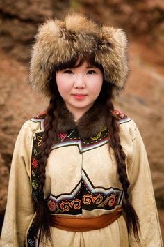 a woman wearing a fur hat and long braids standing in front of some rocks