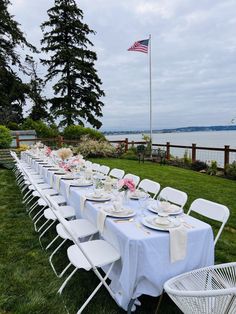 a long table is set with plates and place settings for an outdoor dinner party by the water