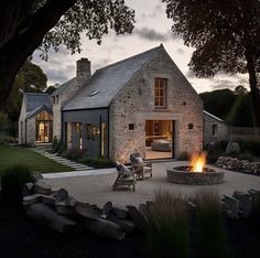 a stone house with a fire pit in the front yard at dusk, surrounded by grass and trees