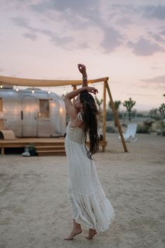 a woman standing in the sand with her arms up and hands behind her head while holding onto a wooden pole