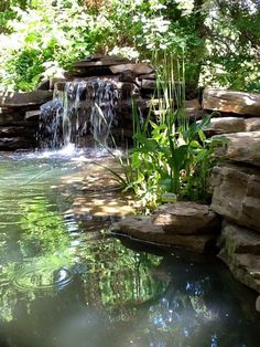 a small waterfall in the middle of a pond surrounded by rocks and plants with water flowing from it