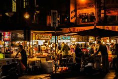 an outdoor market at night with people standing and sitting around the food stalls on the side of the street