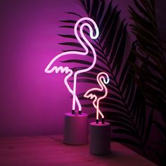 a neon pink flamingo standing next to a palm tree in front of a purple wall