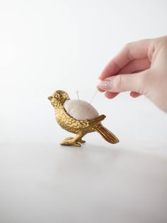 a hand holding a tiny gold bird with needles in it's beak, on a white surface