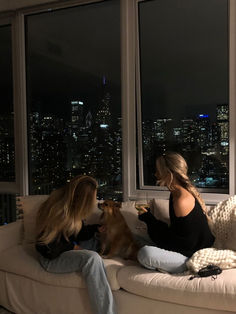 two women sitting on a couch in front of a window with the city lights behind them