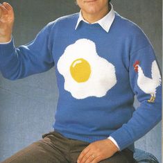 an older man in a blue sweater with fried egg on it's chest, sitting down