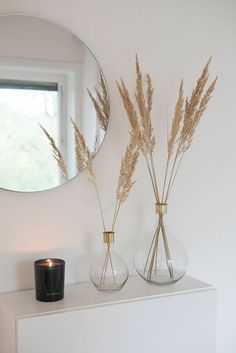 two vases with dried plants sit on a white shelf in front of a round mirror