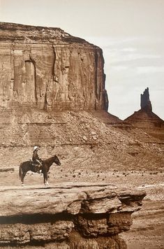 a man riding on the back of a brown horse across a desert field next to a large rock formation