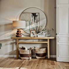 a wooden table topped with baskets under a round mirror next to a wall mounted lamp