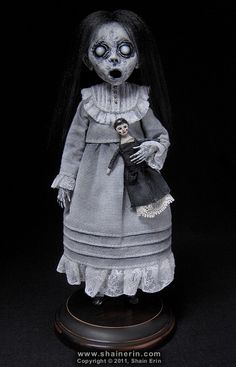 a creepy doll with long black hair wearing a gray dress and holding a small skull