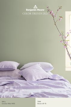 there is a bed with purple sheets and white pillows on the headboard, next to a vase with pink flowers