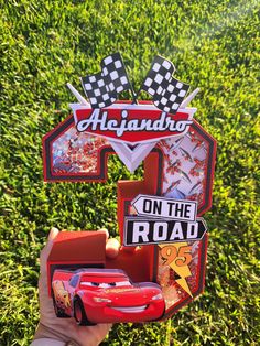 a hand holding up a toy car with cars on the road in front of some grass