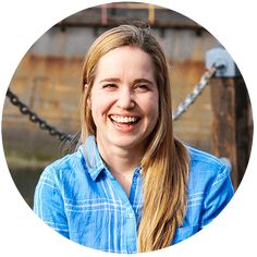 a woman with long blonde hair wearing a blue shirt smiling at the camera in front of a chain link fence