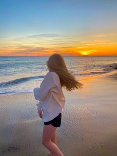 a woman walking on the beach at sunset with her hair blowing in the wind as the sun sets