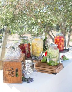 several jars filled with different types of drinks on a white table cloth under a tree