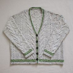 a white knitted sweater with green trims on the front and back, sitting against a wall