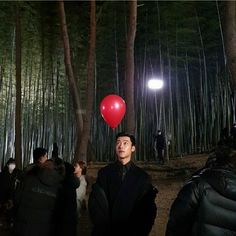 a man with a red balloon on his head standing in the middle of a forest