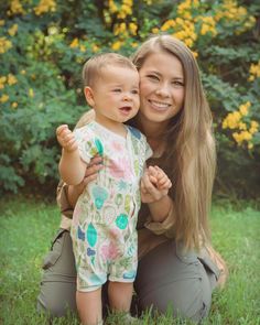 a woman holding a baby in her arms while sitting on the grass with yellow flowers behind her