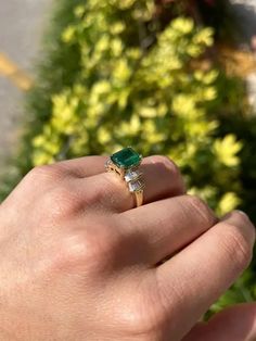 a person holding an emerald and diamond ring in their left hand, with bushes behind them