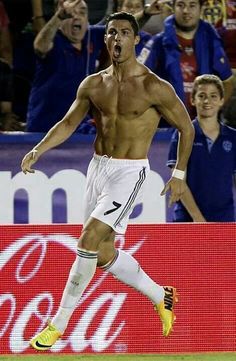 a shirtless male soccer player running on the field with his mouth open and fans in the stands behind him