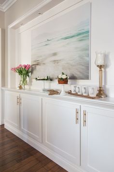 a painting is hanging on the wall next to some white cupboards and vases