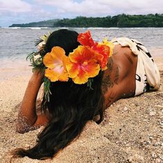 a woman laying on top of a beach next to the ocean with flowers in her hair