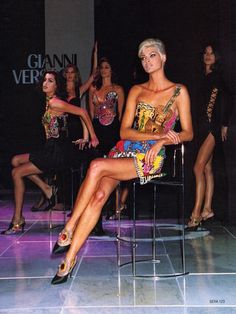 two women sitting on chairs in front of a group of other women wearing dresses and heels