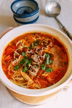 a bowl of soup with noodles, meat and vegetables on a white table cloth next to two spoons