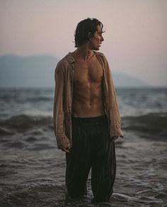 a man standing in the ocean with his shirt off and no shirt on, wearing black pants