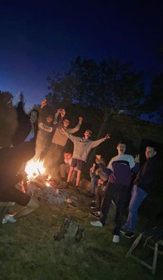 a group of people standing around a campfire at night with their arms in the air