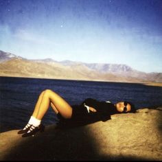 a woman laying on top of a rock next to the ocean with mountains in the background