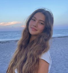 a young woman standing on top of a beach next to the ocean at sunset with her long hair blowing in the wind