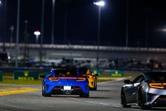 two sports cars driving on a race track at night with lights in the sky behind them