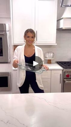 a woman standing in a kitchen holding a glass dish on top of a marble counter