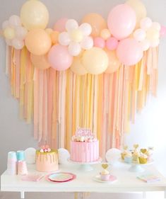 a table topped with cake and balloons next to a wall filled with pink, yellow and white decorations