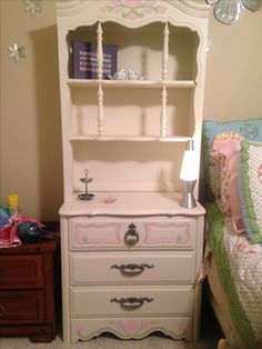 a white dresser sitting next to a bed and a night stand with a lamp on it