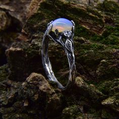 a silver ring with an opal stone in the center on top of mossy rocks