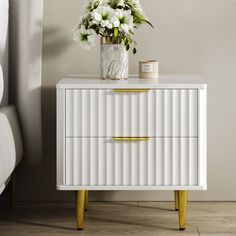 a white dresser with gold handles and flowers in a vase next to the bed side table