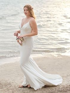 a woman in a wedding dress standing on the beach with her bouquet by the water