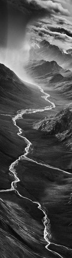 a black and white photo of a river in the middle of some hills with clouds