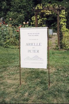 a sign that is in the grass near some bushes and flowers, with an arbor behind it