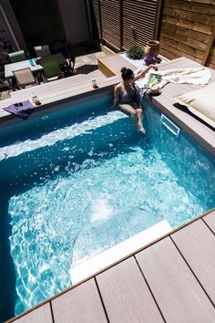 a woman sitting on the edge of a swimming pool next to a deck with lounge chairs