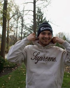 a man in a white hoodie smiles while holding his hands to his ears