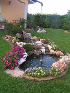 a small pond in the middle of a lawn with flowers and rocks around it, next to a house