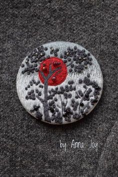 a button with a tree on it sitting on top of a gray sweater, and the words by annna joy written in red