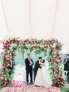 a bride and groom kissing under an archway decorated with pink, red and white flowers