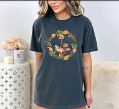 Introducing our charming country style farmhouse fall wreath with mushrooms and moth tshirt! This unique and whimsical tee captures the essence of cozy autumn vibes in an instant. It features an enchanting arrangement of earthy mushrooms and an elegantly detailed moth at the center, adding a touch of nature's beauty to your fall wardrobe. With its rustic farmhouse style and warm color palette, this shirt is the perfect addition to your fall  collection. Enjoy this warm addition to your everyday comfort wear while embracing the spirit of the season with our delightful country style farmhouse fall wreath tshirt. Comfort Colors introduces its garment-dyed t-shirt; a fully customizable tee made 100% with ring-spun cotton. The soft-washed, garment-dyed fabric brings extra coziness to your wardr Vintage Mushroom Print Tops For Fall, Cottagecore Graphic Print Tops For Fall, Fall Cottagecore Tops With Graphic Print, Cottagecore Short Sleeve T-shirt For Fall, Fall Graphic Tee With Mushroom Print, Fall Crew Neck T-shirt With Mushroom Design, Casual Mushroom Design T-shirt For Fall, Fall Mushroom Print Graphic Tee, Casual Fall T-shirt With Mushroom Design