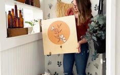 a woman holding up a piece of art in front of a wall with flowers on it